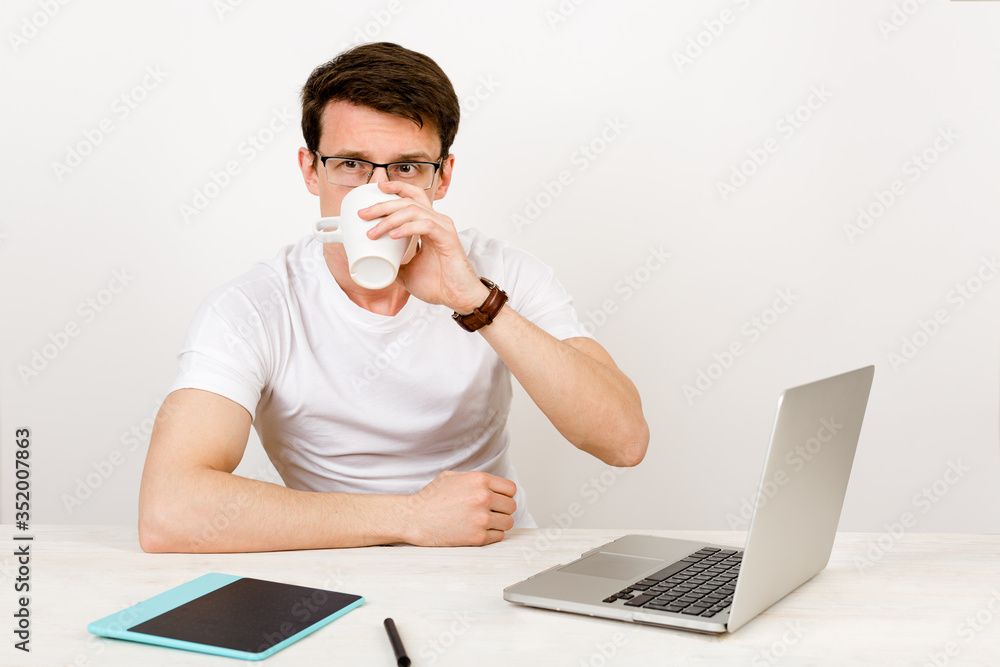 A handsome man with a light smile wearing a T-shirt and glasses sits against a white wall at his desk and work with a laptop. The concept of remote quarantine education and communication A handsome man with a light smile wearing a T-shirt and glasses sits against a white wall at his desk and work with a laptop. The concept of remote quarantine education and communication