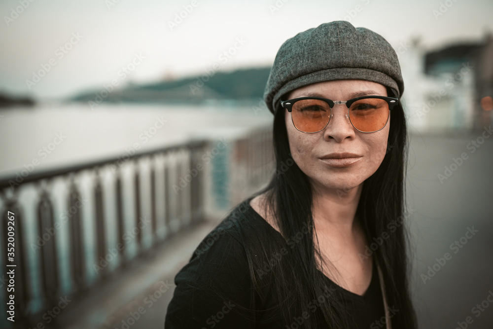 Pretty Asian woman on the river bank. Middle-aged pretty brunette in yellow sunglasses looks at camera on the background of embankment of European city. Portrait. Close up shot