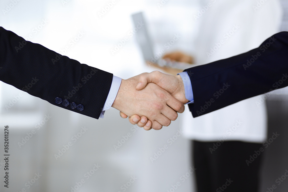 Business people shaking hands at meeting or negotiation, close-up. Group of unknown businessmen and a woman with a laptop stand together in a modern office. Teamwork, partnership and handshake concept