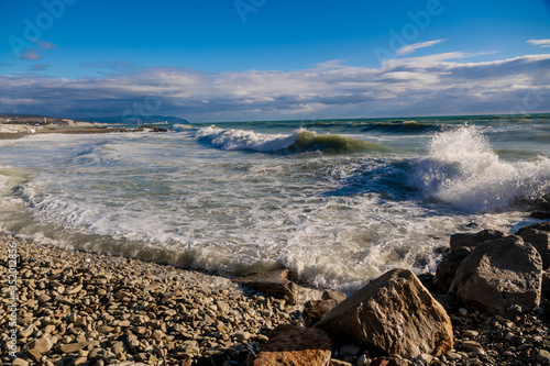 In a storm, waves in snow-white foam fall on the pebble beach of Gelendzhik. In the background Gelendzhik lighthouse and rocks