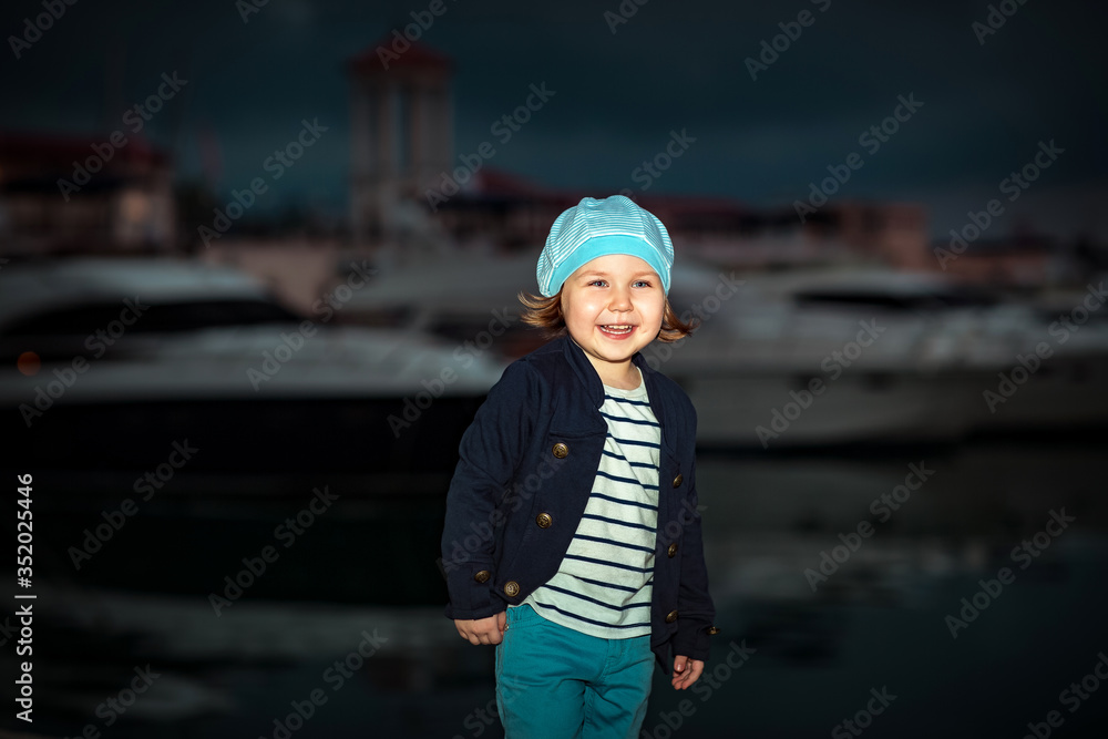 a little beautiful girl, in sea-style clothes, walks in the summer at sunset, on the beach near the yachts
