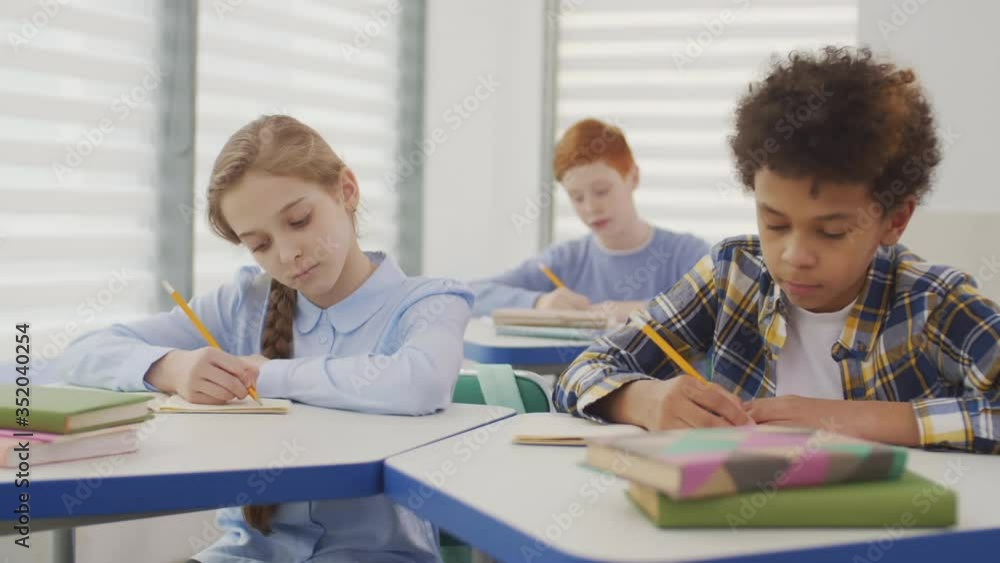 Medium shot of three diverse schoolkids sitting at desks in classroom and doing test. African curly-haired boy coping down from Caucasian girl sitting nearby