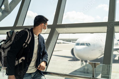 Asian traveler business man wearing face mask waiting to board into airplane, standing in departure terminal in airport. Male passenger traveling by plane transportation during covid19 virus pandemic.