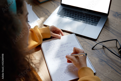 Teen girl college student wear headphones studying from home writing in workbook solving equations learning math sits at desk. Teenage school pupil learn online on laptop, close up over shoulder view. photo