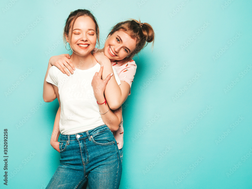 Two young beautiful blond smiling hipster girls in trendy summer hipster jeans clothes. Sexy carefree women posing near blue wall. Trendy and positive models having fun Two young beautiful blond smiling hipster girls in trendy summer hipster jeans clothes. Sexy carefree women posing near blue wall. Trendy and positive models having fun