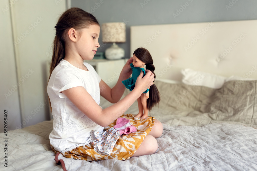 Caucasian girl dresses up doll with long hair, girl plays with dolls on bed in bedroom. Traditional kids games for girls