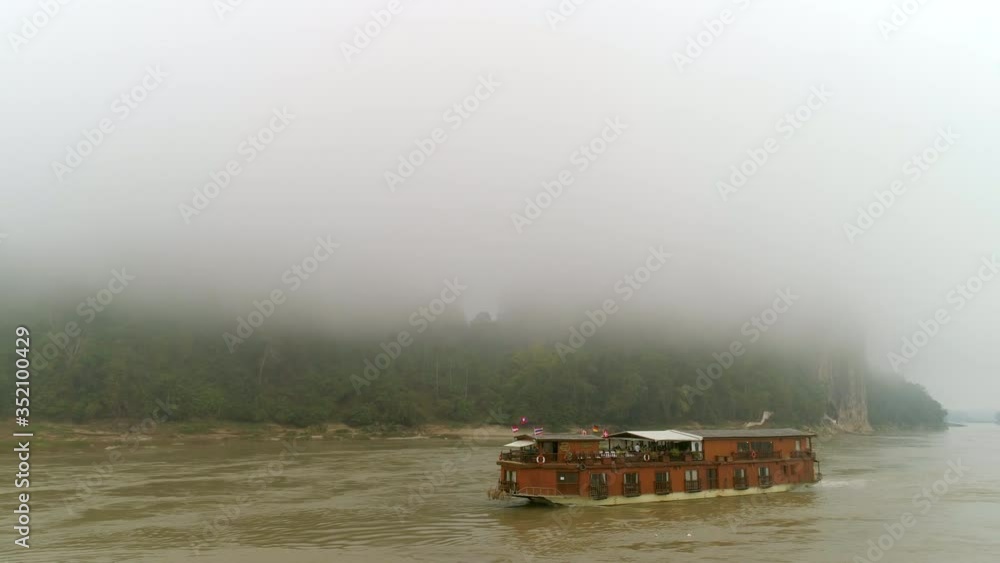 Aerial panning shot of cruise ship moving in river, drone flying over water in foggy weather - Mekong River, Laos Aerial panning shot of cruise ship moving in river, drone flying over water in foggy weather - Mekong River, Laos