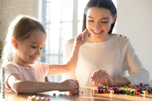 Loving young mother teach excited little daughter string thread wooden beads making stylish bracelets, happy mom or nanny and small preschooler girl child engaged in creative activity or game at home photo