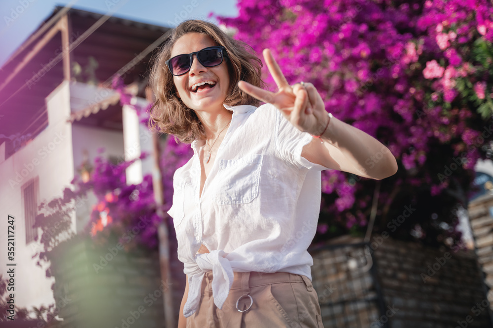 Beautiful young stylish woman traveler in a white shirt in a hat and sunglasses laughs joyfully and shows a victory gesture with her fingers