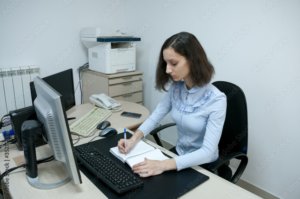 Brunette girl makes notes in a notebook in the office at the computer Brunette girl makes notes in a notebook in the office at the computer