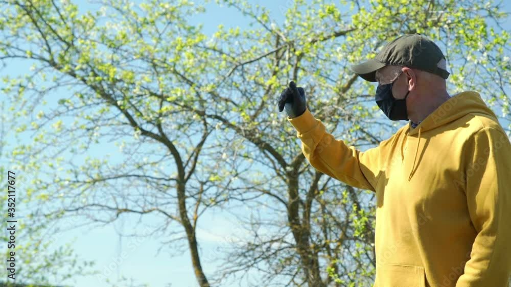 senior man in yellow hoodie black cap and mask waves hand against tree young leaves under blue sky on sunny day slow motion senior man in yellow hoodie black cap and mask waves hand against tree young leaves under blue sky on sunny day slow motion