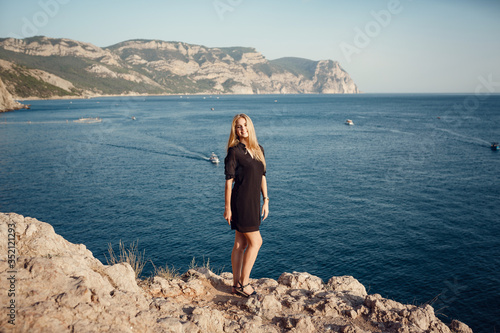 Woman in black dress standing above summer sea on cliff alone.