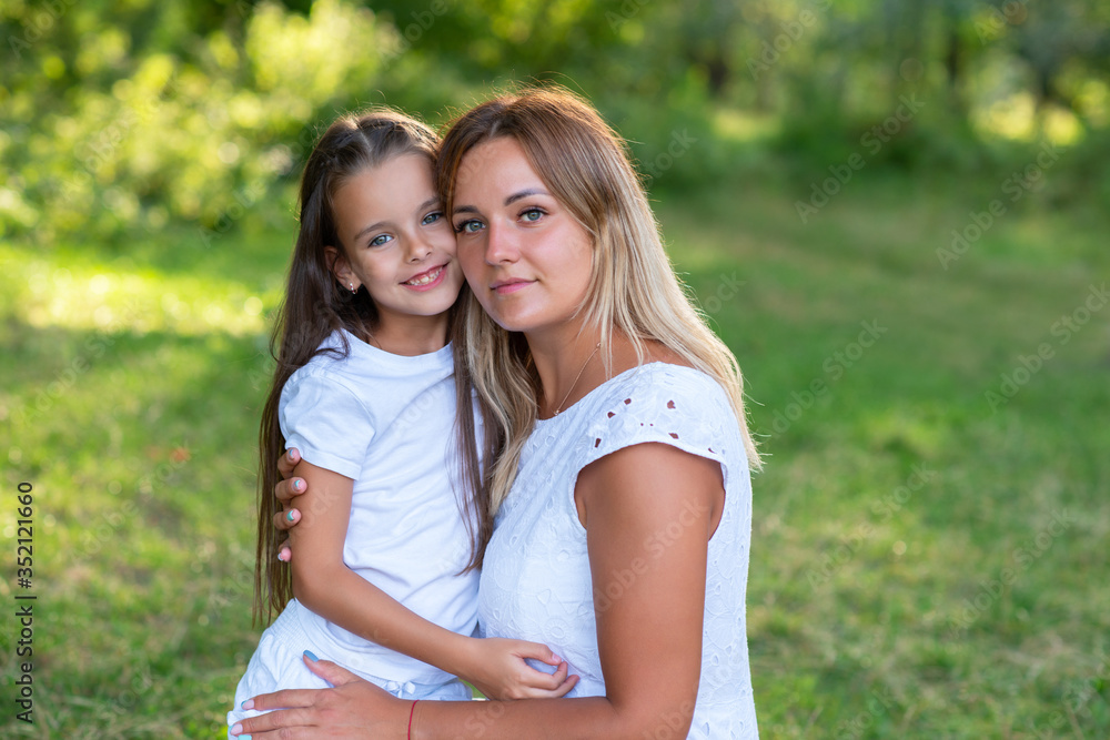 Fototapeta premium Little girl hugs mother, summer nature outdoor. Mom and daughter wearing white clothes against summer greenery. Family walks in park. Trust, kindness, maternity, parenthood, confidence, mother's love