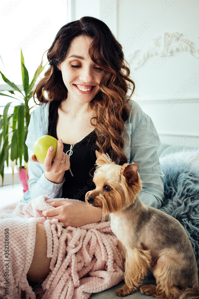 Obraz premium A girl with a curly hair sitting at home on the sofa and holding a green apple on her hand. A Dog. Selective focus. Home plants in the background. Home comfort. Stay home. quarantine.