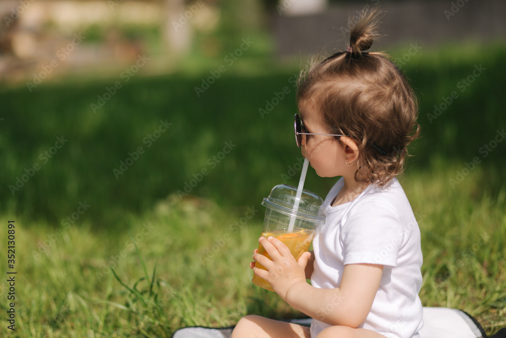 Happy little kid sits outdoors on carpet and drink lemonade. Adorable little girl in white bodysuit Happy little kid sits outdoors on carpet and drink lemonade. Adorable little girl in white bodysuit