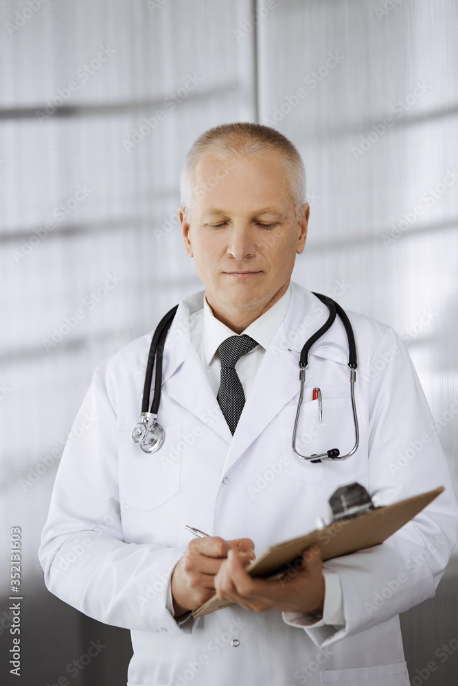 Elderly confident doctor standing straight and using clipboard in clinic. Perfect medical service. Medicine and healthcare concept Elderly confident doctor standing straight and using clipboard in clinic. Perfect medical service. Medicine and healthcare concept