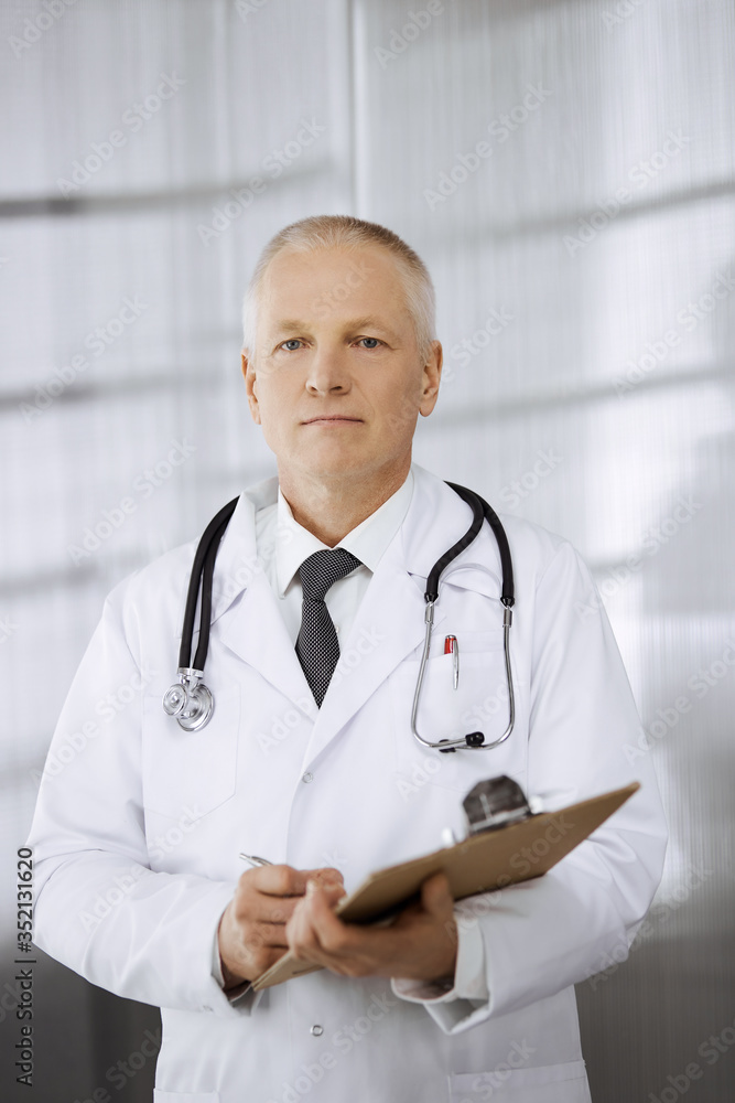 Elderly confident doctor standing straight and using clipboard in clinic. Perfect medical service. Medicine and healthcare concept Elderly confident doctor standing straight and using clipboard in clinic. Perfect medical service. Medicine and healthcare concept