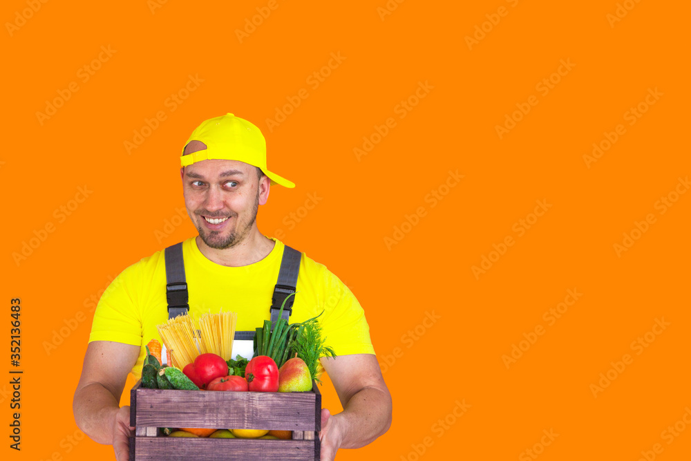 Home delivery food. Wooden box of food, fruits and vegetables for people suffering from coronavirus pandemic consequences, man in a yellow t-shirt on an orange background Home delivery food. Wooden box of food, fruits and vegetables for people suffering from coronavirus pandemic consequences, man in a yellow t-shirt on an orange background