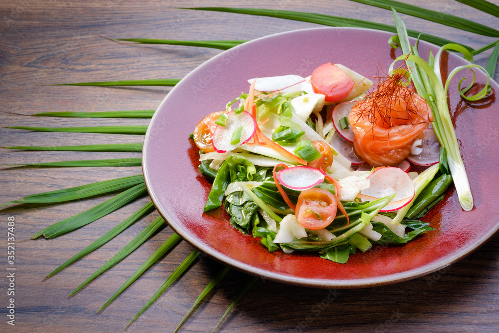 Fresh salmon salad with vegetables on red plate on  wooden table background. Asian salad with fish, onion, carrot, radish, tomato and bok choy. Restaurant menu. Close up, copy space