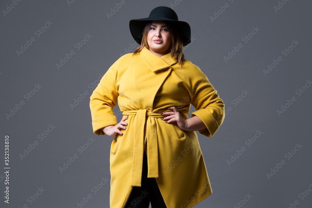 Plus size fashion model in yellow coat and black hat, fat woman on gray background