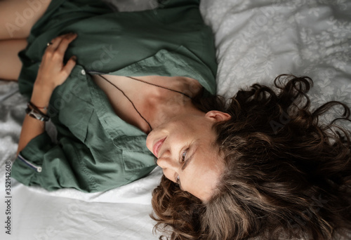 Beautiful charming young curly girl woman in a helen shirt lies on the bed, shot from above.