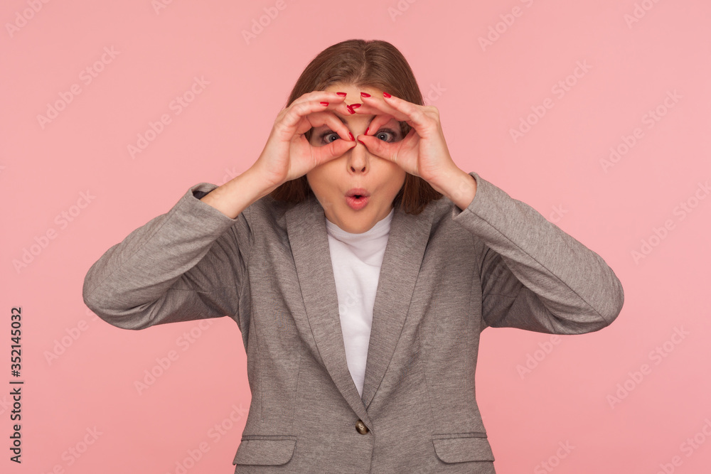 Portrait of surprised young woman in business suit making glasses shape with fingers, looking through binoculars hand gesture and expressing shock, astonishment. indoor studio shot, pink background Portrait of surprised young woman in business suit making glasses shape with fingers, looking through binoculars hand gesture and expressing shock, astonishment. indoor studio shot, pink background