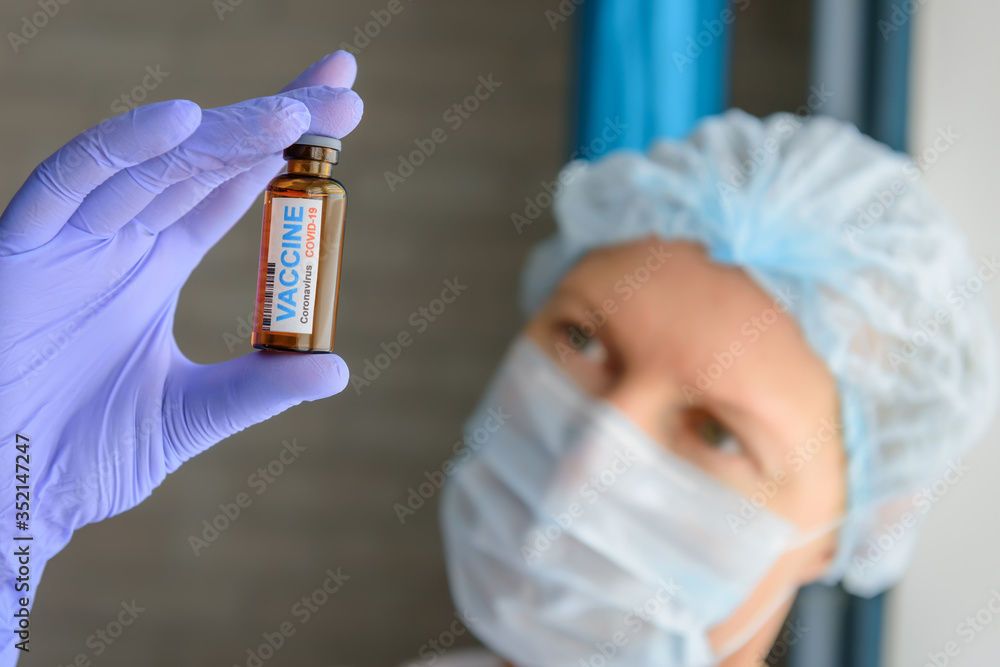 Close up doctor in medical mask and latex gloves holds in her hand vial with covid-19 vaccine. Nurse in white coat  looks at the bottle with coronavirus vaccine, blurred background.