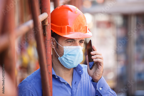 Ingegnere con casco arancione e mascherina Chirurgica lavora in un cantiere in città e parla con il cellulare 