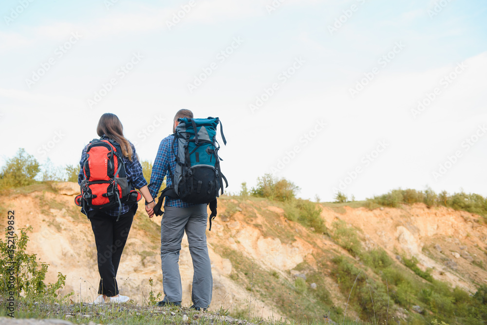 Back view of young pair backpackers with big backpacks holding hands and walking along a road with beautiful mountain landscape on background.