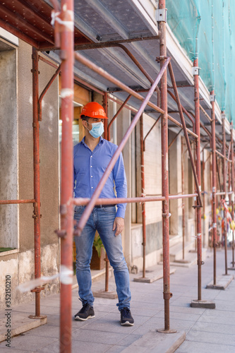 Ingegnere con caschetto arancione e mascherina isolato dentro le transenne di un impalcatura in un cantiere edile