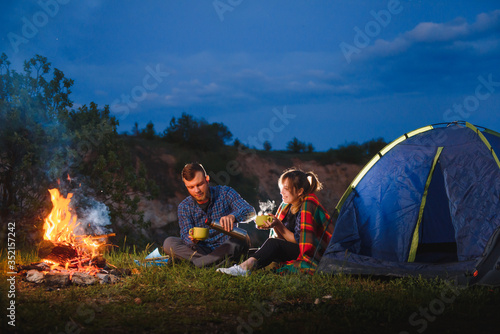 Camping night in mountains. Tourist couple sitting in front of illuminated tent lit by burning campfire. Tourism and outdoor activity concept.