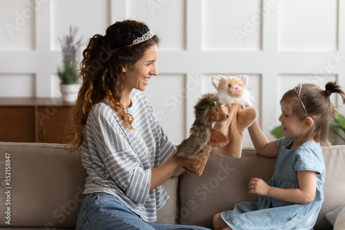 Happy young mother and little daughter wearing princess diadems, having fun with fluffy dolls, sitting on cozy couch, smiling mum and cute preschool girl playing doll theatre game at home