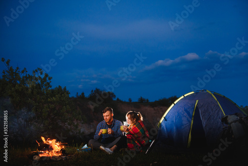 Happy romantic couple travelers resting at bonfire near glowing tourist tent under amazing night sky. The concept of active recreation and travel with a tent