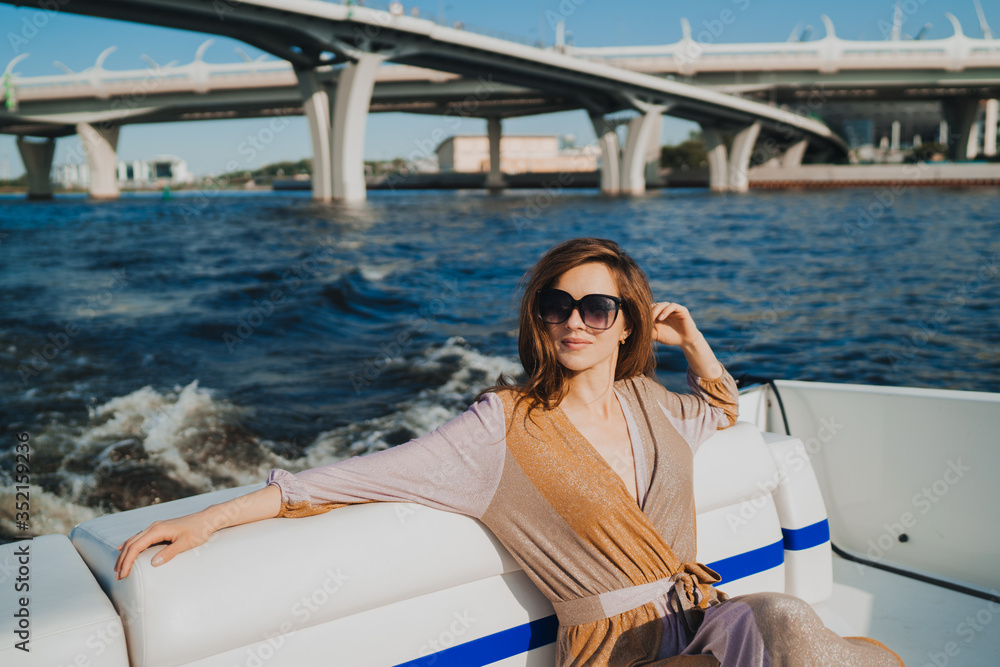 Gorgeous blonde young woman in sunglasses, dressed in pastel dress, enjoying a vacation, satisfied by her life, sitting on  white yacht in the see, bridge on the background. Adventures and vacations.
