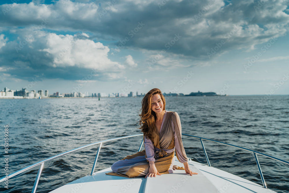 Gorgeous cheerful blonde young woman dressed in pastel dress, enjoying a vacation, sitting on the bow of a white yacht in the see, city on the background and sky with clouds. Adventures and vacations.