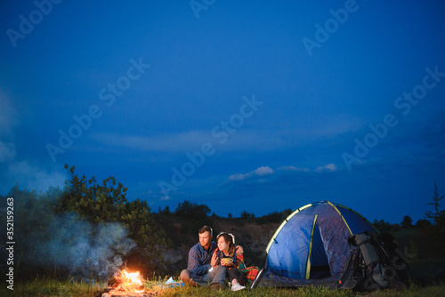 Young couple drinking coffee, and warming up on the campfire in the forest hill in the dusk. The concept of active recreation and travel with a tent