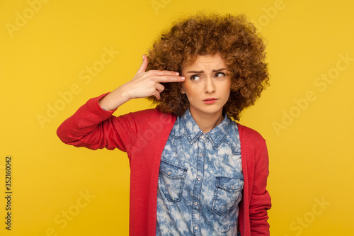 I want to kill myself. Portrait of depressed woman with curly hair pointing finger pistol and shooting head, feeling desperate, thinking over suicide. indoor studio shot isolated on yellow background