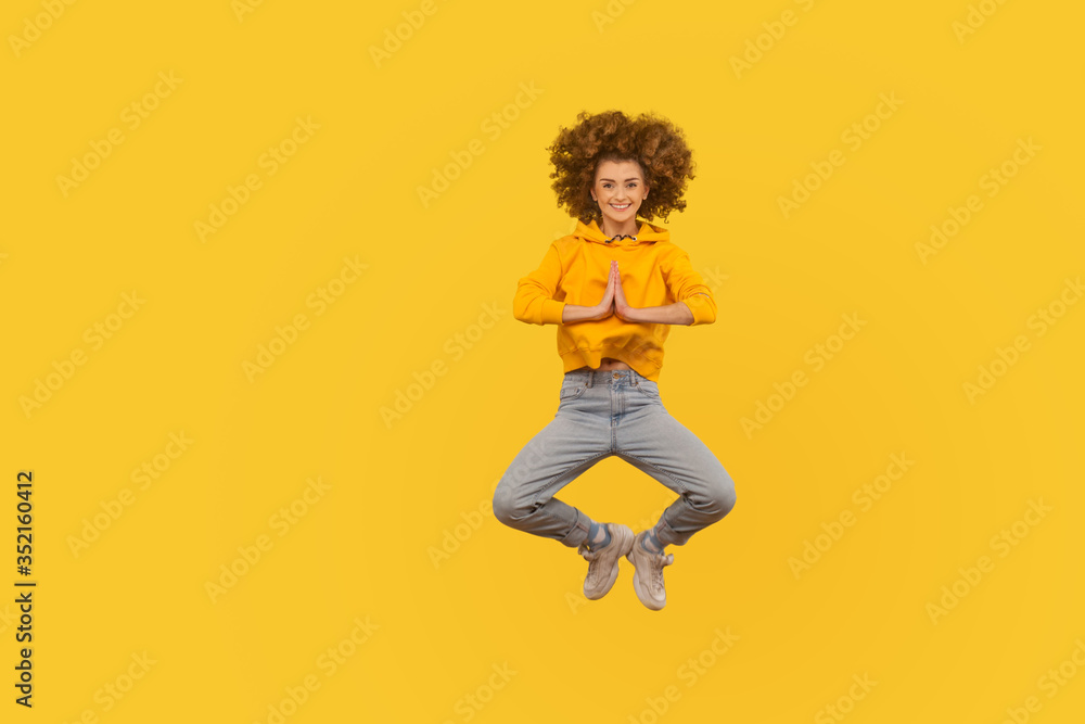 Full length portrait of happy curly-haired woman in urban style outfit flying in air in yoga pose, holding hands namaste gesture and smiling at camera. indoor studio shot isolated on yellow background Full length portrait of happy curly-haired woman in urban style outfit flying in air in yoga pose, holding hands namaste gesture and smiling at camera. indoor studio shot isolated on yellow background