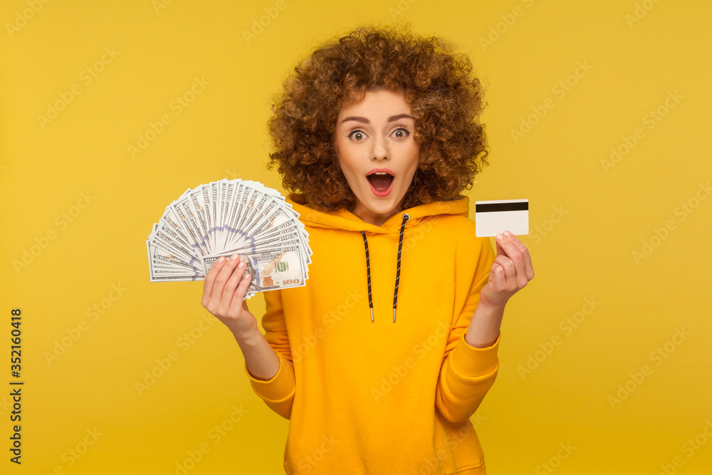 Wow, big money savings, debit account. Portrait of surprised curly-haired girl in urban style hoodie showing dollar banknotes and plastic bank card, looking with amazement. indoor studio shot isolated Wow, big money savings, debit account. Portrait of surprised curly-haired girl in urban style hoodie showing dollar banknotes and plastic bank card, looking with amazement. indoor studio shot isolated