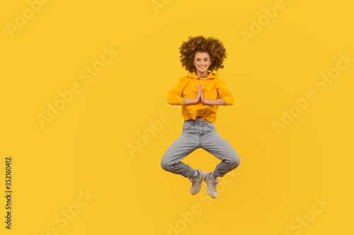 Full length portrait of happy curly-haired woman in urban style outfit flying in air in yoga pose, holding hands namaste gesture and smiling at camera. indoor studio shot isolated on yellow background