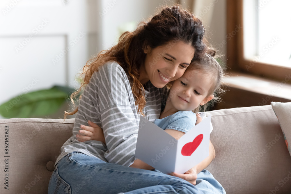 Smiling mother hugging cute little daughter, thanking for gift, sitting on couch at home, preschool girl congratulating mum with birthday or mothers day, presenting greeting card with red heart Smiling mother hugging cute little daughter, thanking for gift, sitting on couch at home, preschool girl congratulating mum with birthday or mothers day, presenting greeting card with red heart