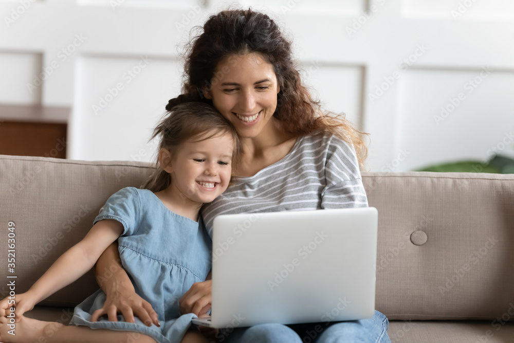 Smiling mother and little daughter looking at laptop screen together, hugging, sitting on cozy couch, spending leisure time at home, watching cartoons, funny video, playing game online Smiling mother and little daughter looking at laptop screen together, hugging, sitting on cozy couch, spending leisure time at home, watching cartoons, funny video, playing game online