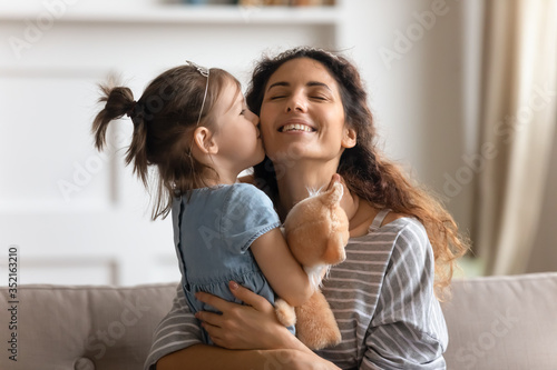 Little girl wearing princess diadem kissing smiling mother on cheek, holding fluffy toy, loving caring young mum and adorable cute daughter hugging, enjoying tender moment, having fun at home