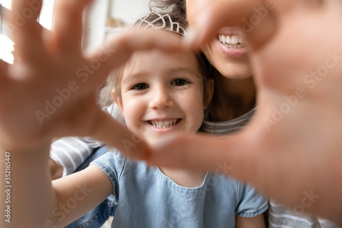 Close up little girl wearing princess diadem and mother showing heart sign with hands, looking through fingers at camera, taking selfie, happy mum and smiling daughter having fun together