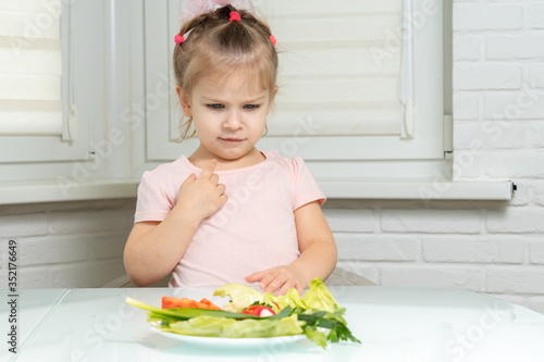 Photos girl preschooler in dislike looks at a plate with vegetables