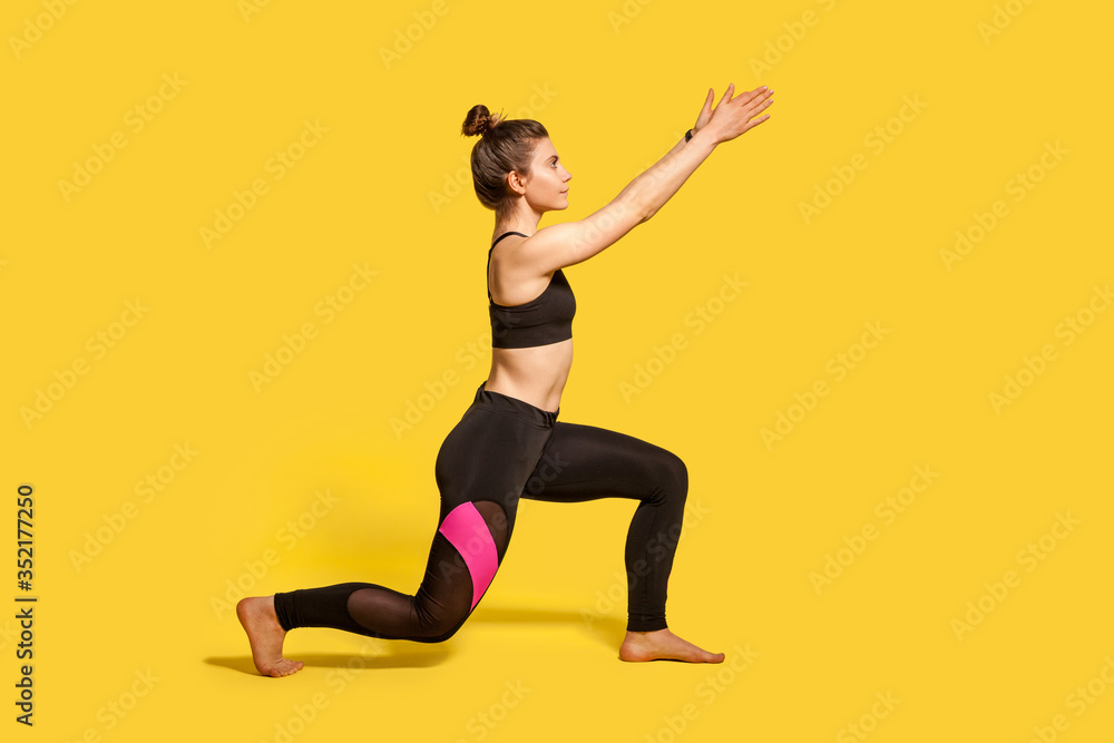 Obraz premium Widok z boku, szczupła młoda kobieta z kok do włosów w obcisłej odzieży sportowej, robi lonży, stojąc na jednym kolanie i unosząc ręce, rozgrzewając mięśnie treningowe. pełnej długości studio strzał, odizolowane na żółto