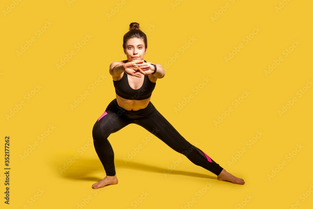 Athletic pretty girl with hair bun in tight sportswear doing sport, lower-body exercise, stretching hands out, warming up training muscles for flexibility. full length studio shot, isolated on yellow