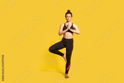 Tree pose. Athletic slim woman with hair bun in tight sportswear practicing yoga, doing Vrksasana exercise on one leg, hands in prayer namaste gesture. full length studio shot, sport workouts isolated © khosrork