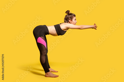 Side view, athletic woman with hair bun in tight sportswear doing sport, bending and stretching hands out, warming up training muscles for flexibility. full length studio shot, isolated on yellow
