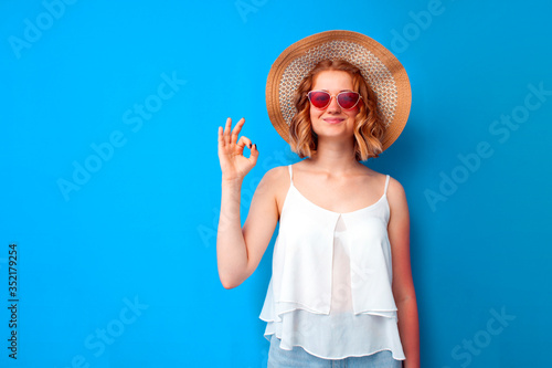 summer concept. girl in a sun hat and glasses shows an okay sign on a blue isolated background, a woman in summer clothes on vacation